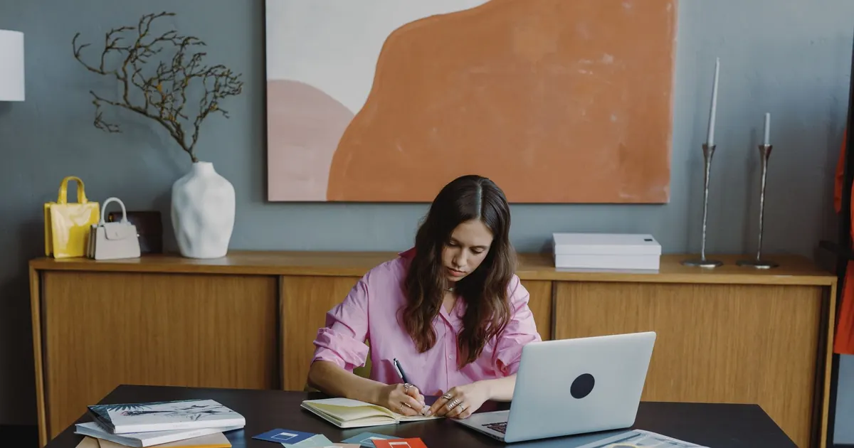 Mulher de camisa rosa sentada à mesa de casa, escrevendo num caderno ao lado do notebook aberto