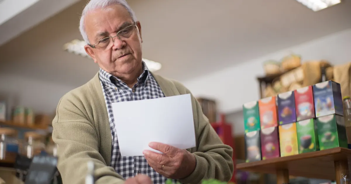 Senhor de cabelo branco e óculos, dono de um pequeno comércio, lendo com atenção uma folha de papel entre as prateleiras da loja