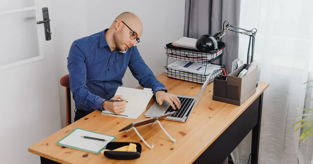 Homem sentado à mesa de casa, falando ao telefone enquanto anota num caderno ao lado do notebook e de uma pilha de papéis