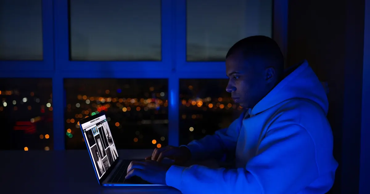 Homem sozinho à noite, de moletom, trabalhando no notebook perto da janela com as luzes da cidade ao fundo